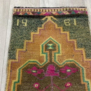 May include: A rectangular woven rug featuring a dark green background with a central stepped geometric pattern in shades of orange and brown. The design includes bright pink floral motifs and the numbers 19 and 81 in yellow at the top corners.