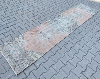May include: A rectangular, vintage-style runner rug with a faded, distressed appearance. The rug features a central peach-colored field with ornate, floral-inspired patterns in shades of blue, gray, and cream along the borders and central design.