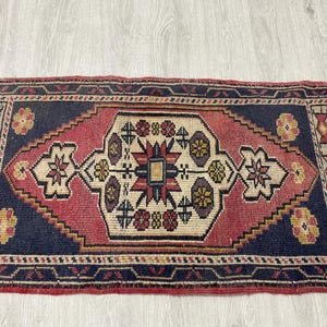 Puede incluir: Una alfombra rectangular con un borde azul oscuro y un medall&oacute;n central en forma de diamante en tonos rojo, crema y morado. El dise&ntilde;o incluye motivos florales geom&eacute;tricos y patrones intrincados distribuidos por todo el campo y los bordes.