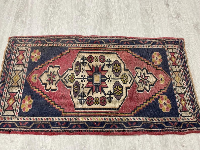 Puede incluir: Una alfombra rectangular con un borde azul oscuro y un medall&oacute;n central en forma de diamante en tonos rojo, crema y morado. El dise&ntilde;o incluye motivos florales geom&eacute;tricos y patrones intrincados distribuidos por todo el campo y los bordes.
