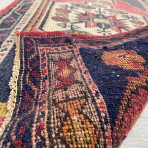 Puede incluir: Una alfombra tejida con un campo azul marino profundo y una esquina doblada que revela un borde rojo y patrones geom&eacute;tricos intrincados en crema, naranja y rojo. El tejido muestra una construcci&oacute;n de pelo denso con bordes anudados visibles.