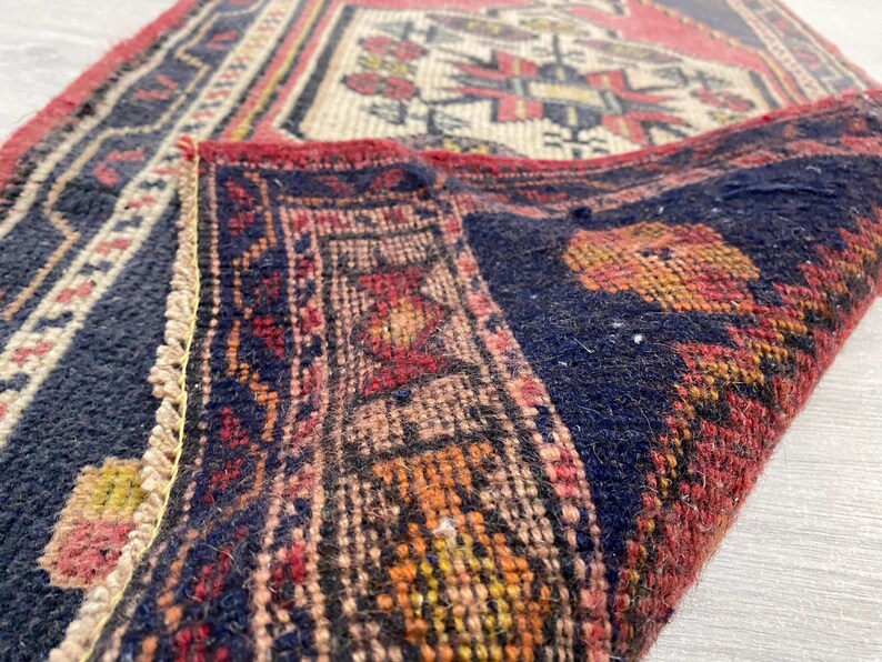 Puede incluir: Una alfombra tejida con un campo azul marino profundo y una esquina doblada que revela un borde rojo y patrones geom&eacute;tricos intrincados en crema, naranja y rojo. El tejido muestra una construcci&oacute;n de pelo denso con bordes anudados visibles.