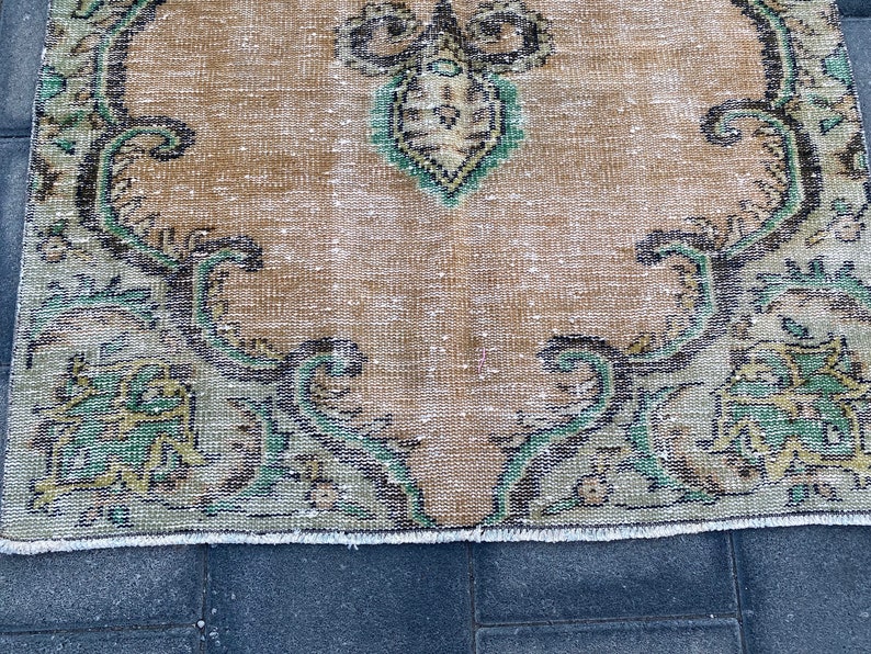 May include: A vintage rug with a faded beige background and a floral pattern in shades of green, black, and gold. The rug has a frayed edge.
