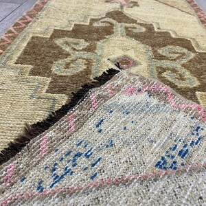May include: A rectangular, hand-woven rug with a beige background and a central brown geometric design. The rug features blue and pink accents, with a pink border and fringed edges. The rug's design is intricate and traditional.