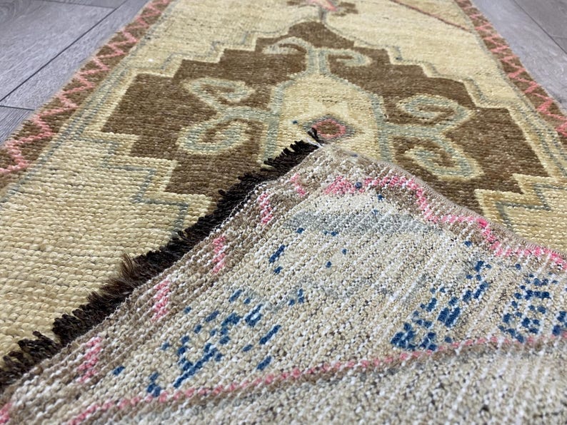 May include: A rectangular, hand-woven rug with a beige background and a central brown geometric design. The rug features blue and pink accents, with a pink border and fringed edges. The rug's design is intricate and traditional.