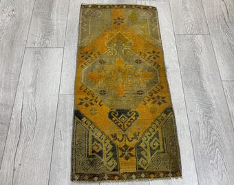 Puede incluir: Una alfombra rectangular de estilo vintage con un patrón central de diamantes en tonos dorados y verde oliva. La alfombra presenta intrincados diseños geométricos y motivos florales, con un borde oscuro. La alfombra está colocada sobre un suelo de madera gris claro.