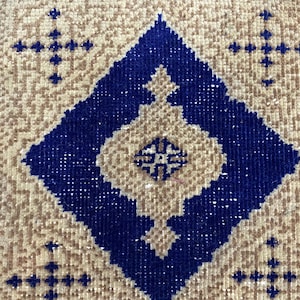 Op de afbeelding: Een close-up van een beige en blauw gepatroneerd tapijt met een ruitvormig ontwerp. Het tapijt heeft een franje rand en is gemaakt van een zacht, geweven materiaal.