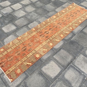 Puede incluir: Una alfombra larga y rectangular con un diseño persa tradicional. La alfombra tiene un fondo rojo con un patrón beige y marrón. La alfombra está sobre un suelo de baldosas grises.