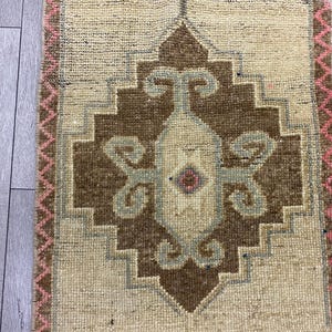 May include: A rectangular rug with a central geometric design in brown, beige, and light blue. The rug has a pink zig-zag border. The rug is made of woven material and is likely used as a decorative floor covering.