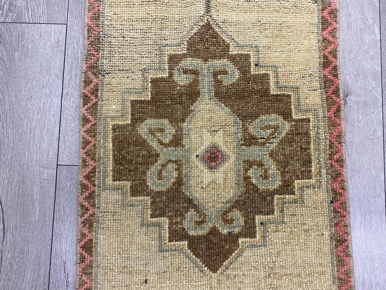 May include: A rectangular rug with a central geometric design in brown, beige, and light blue. The rug has a pink zig-zag border. The rug is made of woven material and is likely used as a decorative floor covering.