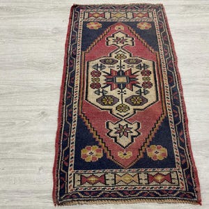 Puede incluir: Una alfombra tejida rectangular que presenta un motivo central de diamante en tonos rojo, azul marino y crema. El dise&ntilde;o incluye patrones florales geom&eacute;tricos y un borde decorativo con formas repetitivas en amarillo, rojo y azul en la pieza.