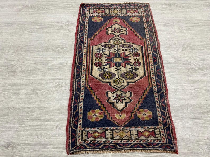 Puede incluir: Una alfombra tejida rectangular que presenta un motivo central de diamante en tonos rojo, azul marino y crema. El dise&ntilde;o incluye patrones florales geom&eacute;tricos y un borde decorativo con formas repetitivas en amarillo, rojo y azul en la pieza.