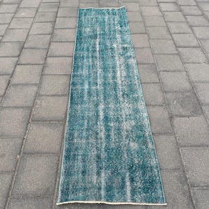 May include: A long, rectangular runner rug featuring a distressed, faded teal and white pattern. The textile displays a weathered appearance with visible variations in color density across its surface, creating a textured, worn-in aesthetic.