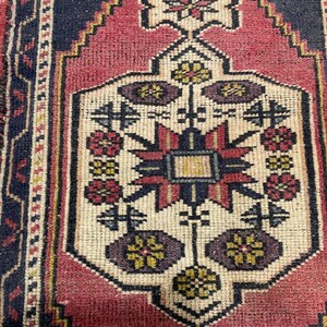 Puede incluir: Una alfombra rectangular con un medall&oacute;n central de color crema que presenta un patr&oacute;n geom&eacute;trico de estrella en rojo, azul oscuro y amarillo. El medall&oacute;n est&aacute; rodeado por un campo rojo intenso y un borde azul oscuro con motivos geom&eacute;tricos.