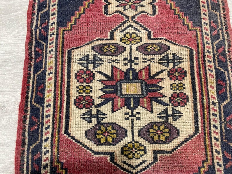 Puede incluir: Una alfombra rectangular con un medall&oacute;n central de color crema que presenta un patr&oacute;n geom&eacute;trico de estrella en rojo, azul oscuro y amarillo. El medall&oacute;n est&aacute; rodeado por un campo rojo intenso y un borde azul oscuro con motivos geom&eacute;tricos.
