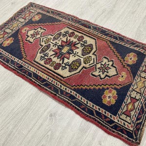 Puede incluir: Una alfombra rectangular con fondo azul marino oscuro y un medall&oacute;n geom&eacute;trico central en tonos rojo apagado y crema. El dise&ntilde;o incluye motivos florales complejos en amarillo, rojo y azul, con bordes terminados en flecos en los lados cortos.