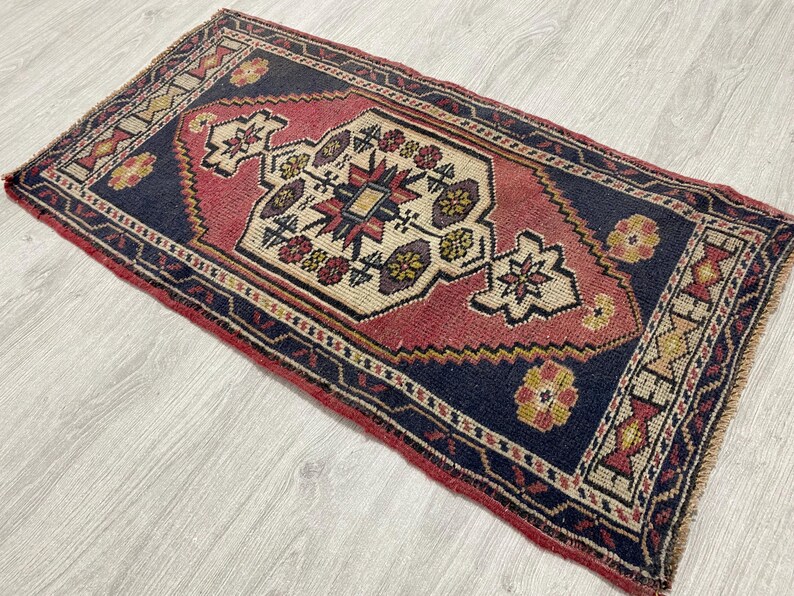 Puede incluir: Una alfombra rectangular con fondo azul marino oscuro y un medall&oacute;n geom&eacute;trico central en tonos rojo apagado y crema. El dise&ntilde;o incluye motivos florales complejos en amarillo, rojo y azul, con bordes terminados en flecos en los lados cortos.