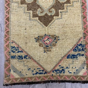 May include: A small, rectangular rug with a beige central field. The rug features a geometric design in brown, blue, and pink. The border is a combination of pink and brown, with a fringe at the bottom. The rug is likely handmade.