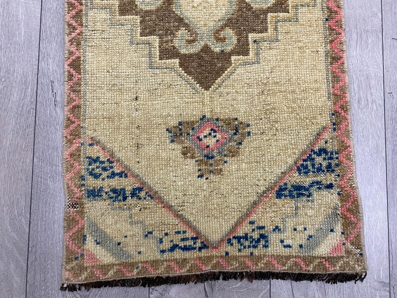 May include: A small, rectangular rug with a beige central field. The rug features a geometric design in brown, blue, and pink. The border is a combination of pink and brown, with a fringe at the bottom. The rug is likely handmade.