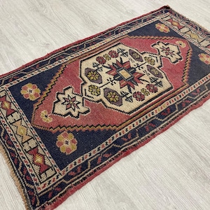 Puede incluir: Una alfombra rectangular con un medall&oacute;n central en forma de diamante de color crema, rodeado por un campo azul marino profundo y un borde rojo. El dise&ntilde;o incluye motivos florales geom&eacute;tricos en amarillo, morado y rojo, con flecos.