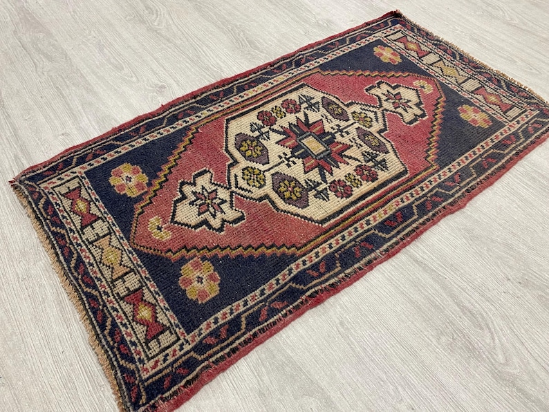 Puede incluir: Una alfombra rectangular con un medall&oacute;n central en forma de diamante de color crema, rodeado por un campo azul marino profundo y un borde rojo. El dise&ntilde;o incluye motivos florales geom&eacute;tricos en amarillo, morado y rojo, con flecos.