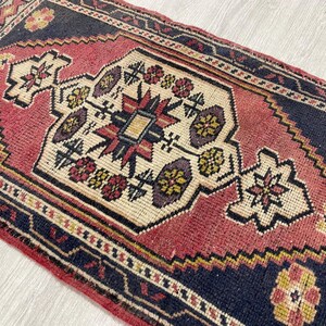 Puede incluir: Una alfombra rectangular que presenta un medall&oacute;n central de color crema con motivos geom&eacute;tricos y florales en rojo, amarillo y azul oscuro. La alfombra tiene un campo rojo intenso rodeado por un borde azul oscuro con patrones complejos.