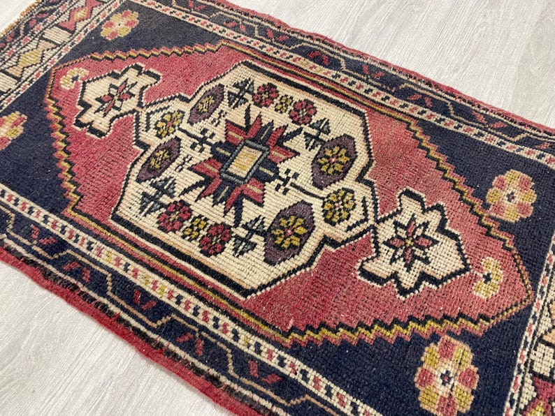 Puede incluir: Una alfombra rectangular que presenta un medall&oacute;n central de color crema con motivos geom&eacute;tricos y florales en rojo, amarillo y azul oscuro. La alfombra tiene un campo rojo intenso rodeado por un borde azul oscuro con patrones complejos.
