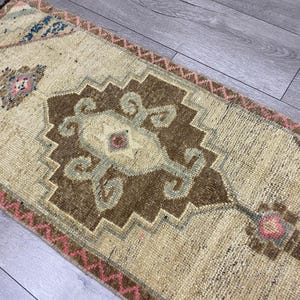 May include: A rectangular, beige rug with a central brown geometric design and accents of blue and pink. The rug features a detailed pattern and a fringed edge, suitable for home decor.