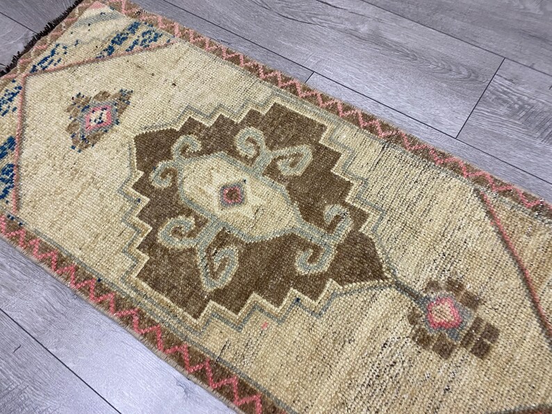 May include: A rectangular, beige rug with a central brown geometric design and accents of blue and pink. The rug features a detailed pattern and a fringed edge, suitable for home decor.