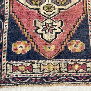 Puede incluir: Una alfombra tejida rectangular con un motivo central de diamante en tonos rojo apagado, crema y azul oscuro. El dise&ntilde;o incluye patrones florales geom&eacute;tricos en amarillo y morado, enmarcados por un borde azul oscuro con detalles decorativos en rojo y crema.