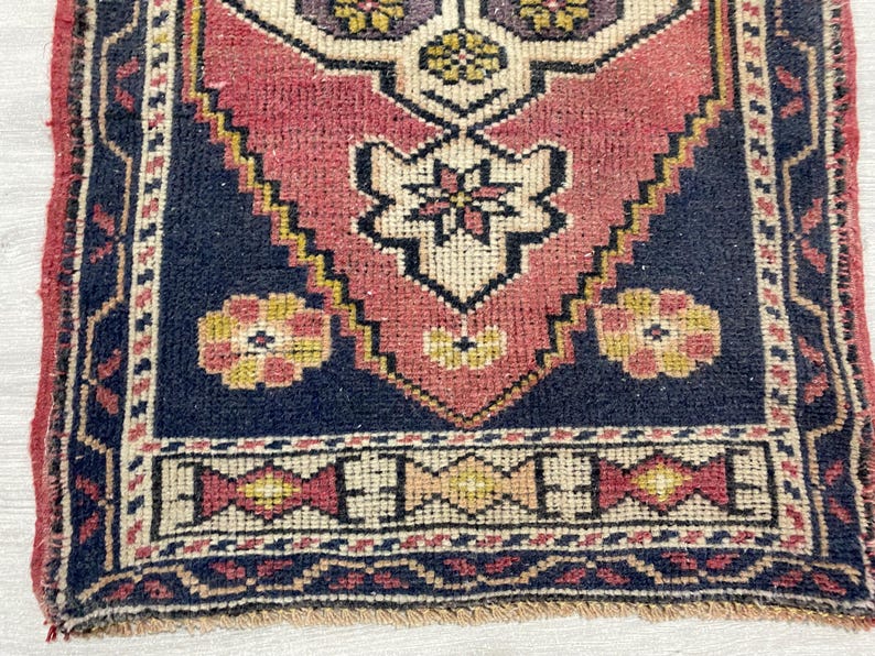 Puede incluir: Una alfombra tejida rectangular con un motivo central de diamante en tonos rojo apagado, crema y azul oscuro. El dise&ntilde;o incluye patrones florales geom&eacute;tricos en amarillo y morado, enmarcados por un borde azul oscuro con detalles decorativos en rojo y crema.