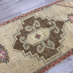 May include: A rectangular, beige rug with a central brown geometric design and accents of pink, blue, and light green. The rug has a textured appearance and a decorative border.