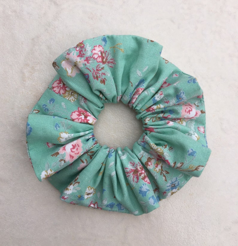 May include: A green fabric scrunchie with a floral pattern. The scrunchie is made of a soft, stretchy fabric and is perfect for holding hair in place.