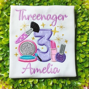 May include: White t-shirt with the embroidered text "Threenager" and "Amelia". The design includes a makeup brush, a compact with glitter, a number 3, lipstick, and nail polish. Gold star accents complete the design.