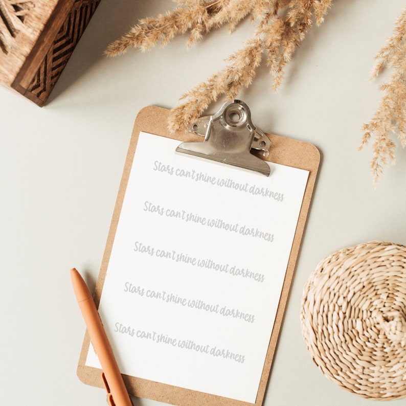 May include: A clipboard with a white sheet of paper and a brown wooden frame. The paper has the text "Stars can't shine without darkness" written five times in cursive handwriting. A brown pen is lying on the clipboard.
