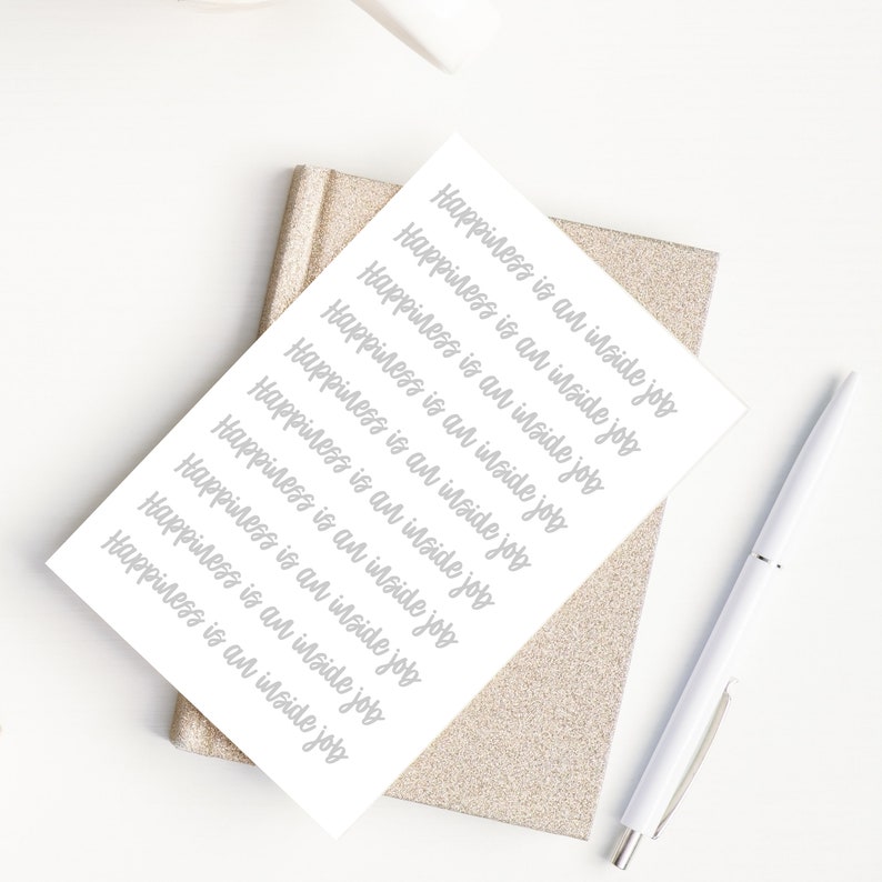 Puede incluir: Una tarjeta blanca con la frase "Happiness is an inside job" repetida 10 veces en escritura cursiva gris. La tarjeta est&aacute; sobre un cuaderno de brillantina dorada con un bol&iacute;grafo blanco al lado.