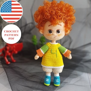 May include: Crochet pattern PDF for a red-haired boy doll wearing a yellow shirt with green and orange stripes, white shorts, and blue shoes.