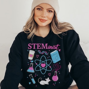 Pu&ograve; includere: Felpa nera con la scritta "STEMinist" in rosa e viola. Il design include grafiche a tema scientifico come una tazza di caff&egrave;, un filamento di DNA, un atomo, un libro, provette e un topo.