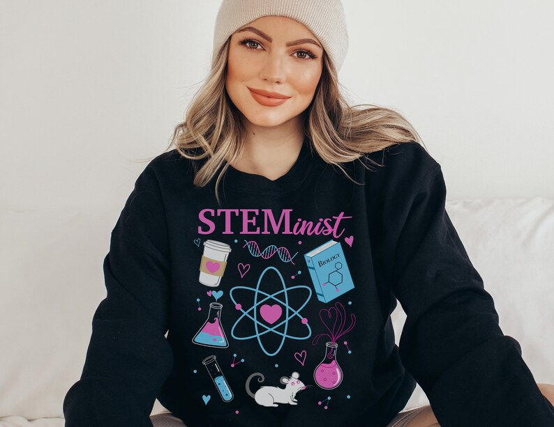 Pu&ograve; includere: Felpa nera con la scritta "STEMinist" in rosa e viola. Il design include grafiche a tema scientifico come una tazza di caff&egrave;, un filamento di DNA, un atomo, un libro, provette e un topo.