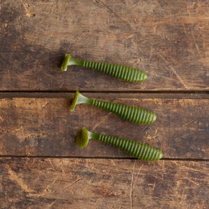 May include: Three green soft plastic fishing lures with ribbed bodies and paddle tails are arranged on a weathered wooden surface. The lures are a vibrant green color, and the ribbed design adds texture. The wooden surface has a rustic, aged appearance.