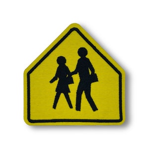 Puede incluir: Parche bordado amarillo y negro con una señal de cruce escolar. La señal representa a dos niños caminando, uno de ellos lleva una mochila.