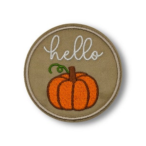 Puede incluir: Un parche redondo de tela beige con un "hola" bordado en blanco y una calabaza bordada en marrón con un tallo verde.