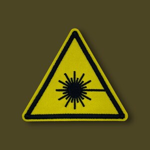 May include: Yellow triangle warning sign with a black border and a black laser beam symbol inside. The sign is made of fabric and has a stitched border.