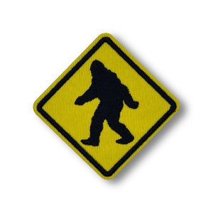 May include: Yellow diamond-shaped warning sign with a black silhouette of Bigfoot walking. The sign is made of fabric and has a stitched border.