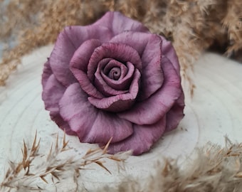 May include: A single, large, purple rose made of polymer clay. The rose is sitting on a light brown, wooden surface.