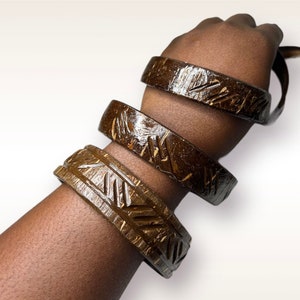 May include: Three brown coconut shell bracelets with intricate carved designs. The bracelets are stacked on a person's arm.