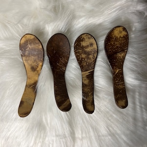 May include: Four brown coconut shell spoons with a natural wood grain pattern. The spoons are arranged in a row on a white fluffy surface.