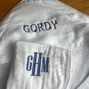 Może przedstawiać: Biały ręcznik z kapturem dla dziecka z imieniem "GORDY" wyhaftowanym na niebiesko z czerwonymi gwiazdkami. Pasująca myjka ma wyhaftowane inicjały "GHM" w kolorze niebieskim. Ręcznik wykonany jest z miękkiej, teksturowanej tkaniny.