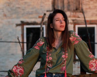Puede incluir: Una mujer con una blusa verde bordada con detalles florales rosas y pantalones vaqueros azules, de pie frente a un edificio rústico.