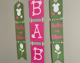 Op de afbeelding: Een roze en groene baby shower banner met het woord "BABY" in grote roze letters en de zin "Baby in Bloom" aan elke kant. De banner is versierd met bloemen en een klein babypakje.